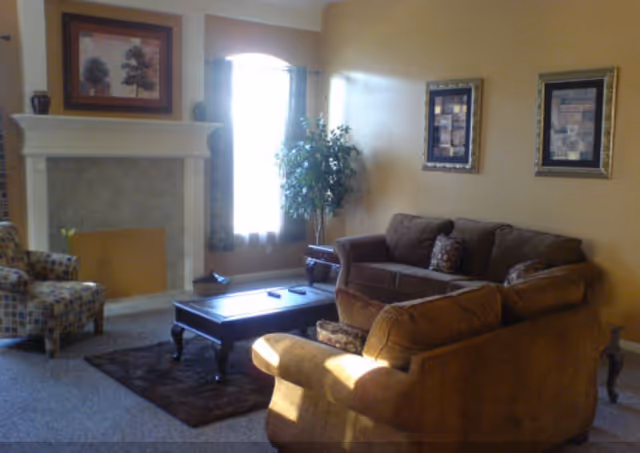 A cozy living room with a brown sofa, a patterned armchair, a wooden coffee table on a dark rug, a fireplace with a framed picture above it, and a window with curtains letting in natural light. There are two framed artworks on the wall and a potted plant near the window.