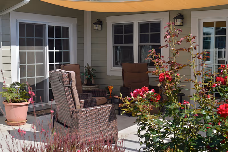 Outdoor patio area with cushioned wicker chairs arranged around a small table, surrounded by flowering plants and shrubs, adjacent to a building with sliding glass doors and windows.