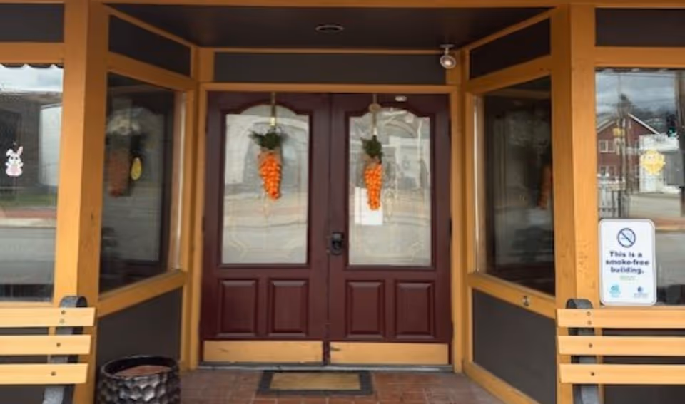 Double wooden doors framed by orange-trim porch, benches outside with hanging orange decorations.