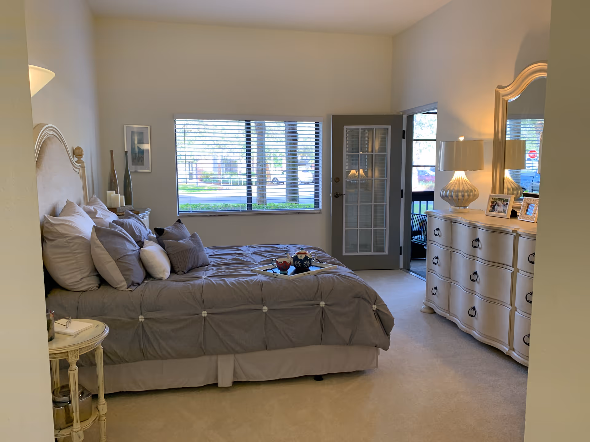 A cozy bedroom with a large bed covered in gray bedding and multiple pillows. There is a small round side table next to the bed with a glass and a small item on it. A dresser with a large mirror, a lamp, and framed photos is on the right side of the room. A window with blinds and a glass door leading outside are visible in the background, letting in natural light.