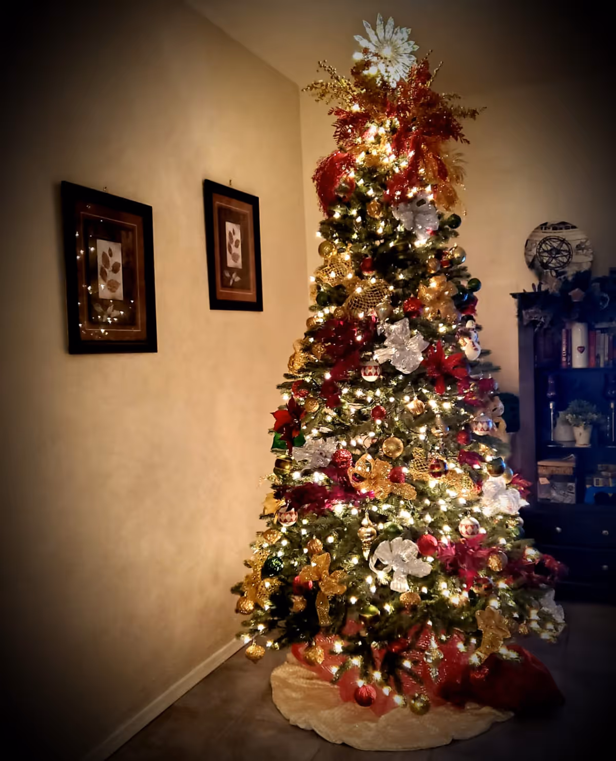 A beautifully decorated Christmas tree adorned with white lights, gold and red ornaments, ribbons, and bows stands in a cozy living room corner. Two framed pictures hang on the adjacent wall, and a dark wooden bookshelf with various items is visible in the background.