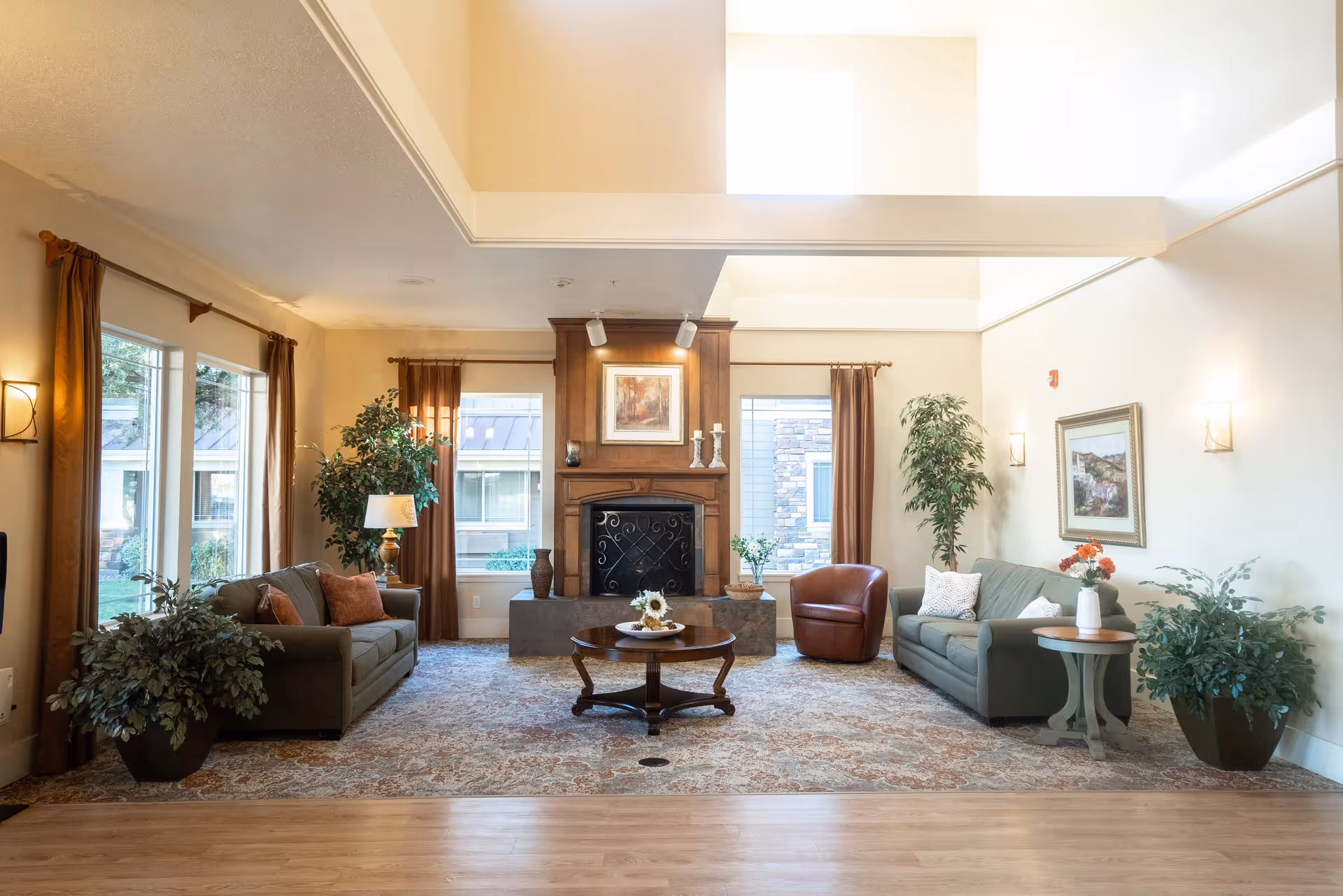 A bright and spacious living room with two green sofas, a brown leather armchair, a wooden coffee table, and a fireplace with a framed picture above it. The room has large windows with brown curtains, several potted plants, wall sconces, and a patterned area rug over a wooden floor.