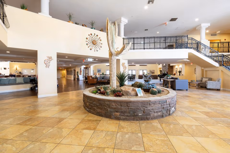 Spacious senior living facility lobby with a large circular stone planter in the center containing a tall cactus and other desert plants. The area features tiled flooring, beige walls, and a mezzanine with black railing. Comfortable seating areas with sofas and chairs are visible in the background, along with decorative wall art and potted plants.