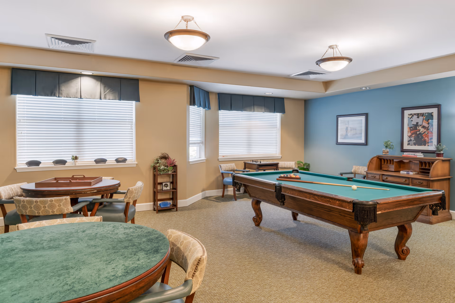 A recreational room with a pool table in the center, surrounded by chairs and tables. The room has beige walls with one blue accent wall, two windows with blinds and valances, and ceiling lights. There is a wooden roll-top desk against the blue wall with framed artwork above it, and a small shelving unit with plants and decor near the windows.