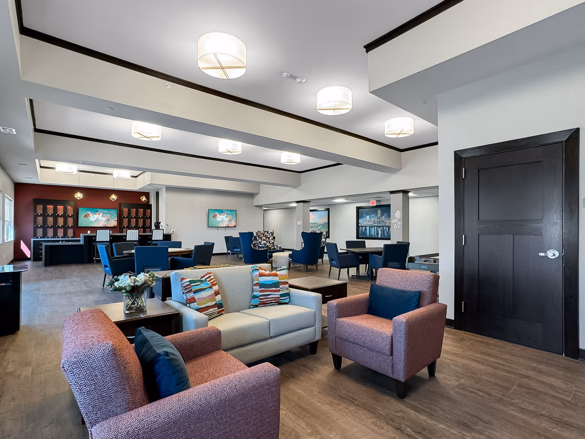 A spacious and well-lit common area in a senior living facility featuring a seating arrangement with a beige sofa and two pink armchairs with colorful cushions. The room has multiple tables and chairs, modern ceiling lights, and artwork on the walls. The floor is wooden, and there is a dark wooden door on the right side.