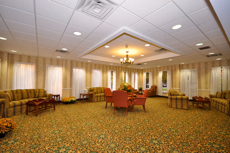 A spacious, carpeted senior living lounge with couches, chairs, and a central chandelier.