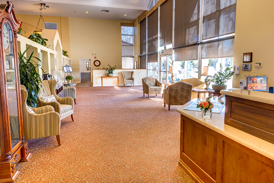 A spacious and well-lit assisted living facility common area with large windows covered by roller shades allowing natural light to fill the room. The space features comfortable armchairs, a wooden reception desk with a small flower arrangement, a tall grandfather clock, and various plants placed around the room. The carpet is patterned in warm tones, and the walls are painted a soft beige color.