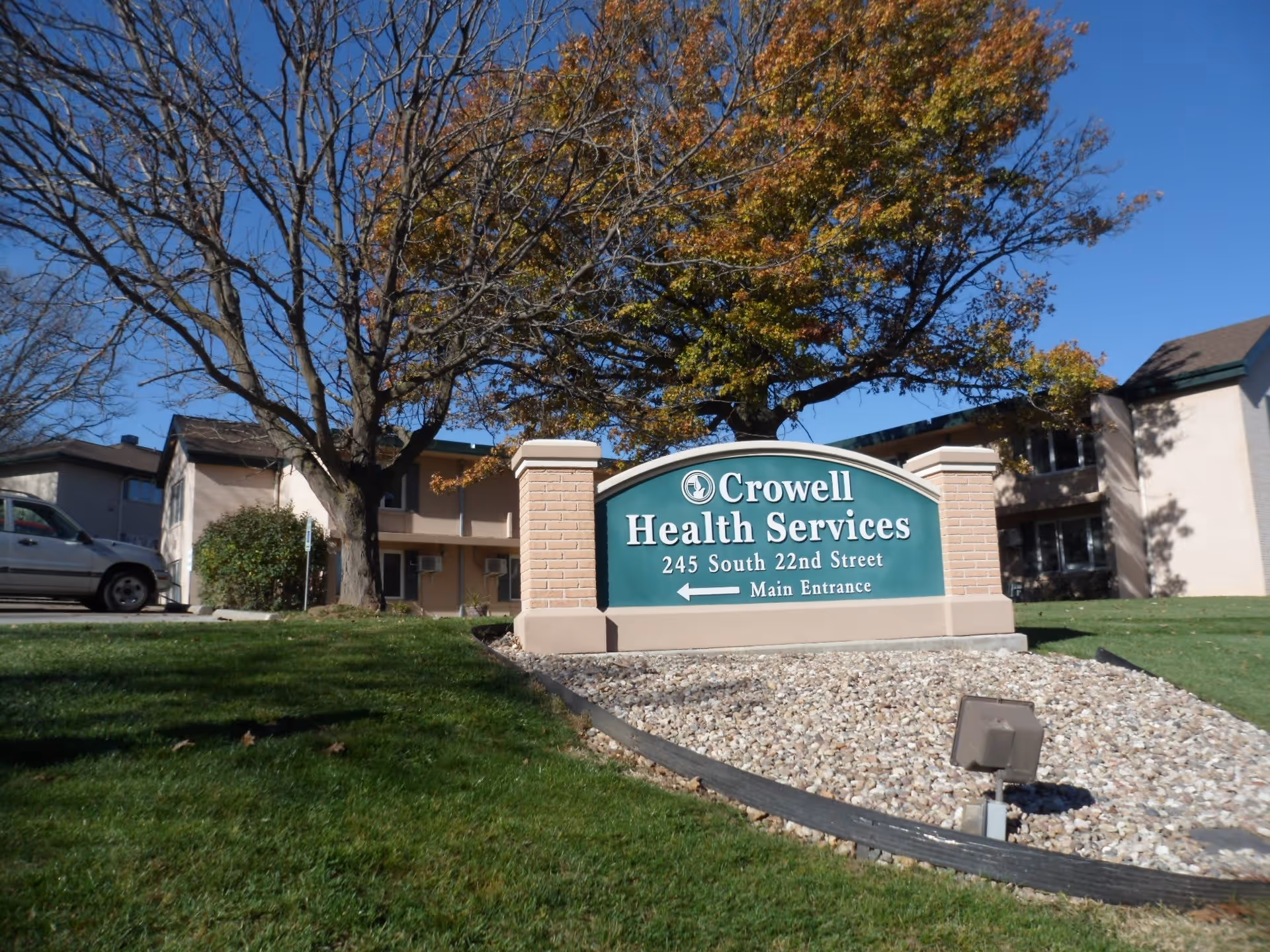 Outdoor view of Crowell Health Services building with a green sign displaying the facility name and address, surrounded by grass, trees with autumn leaves, and a clear blue sky.