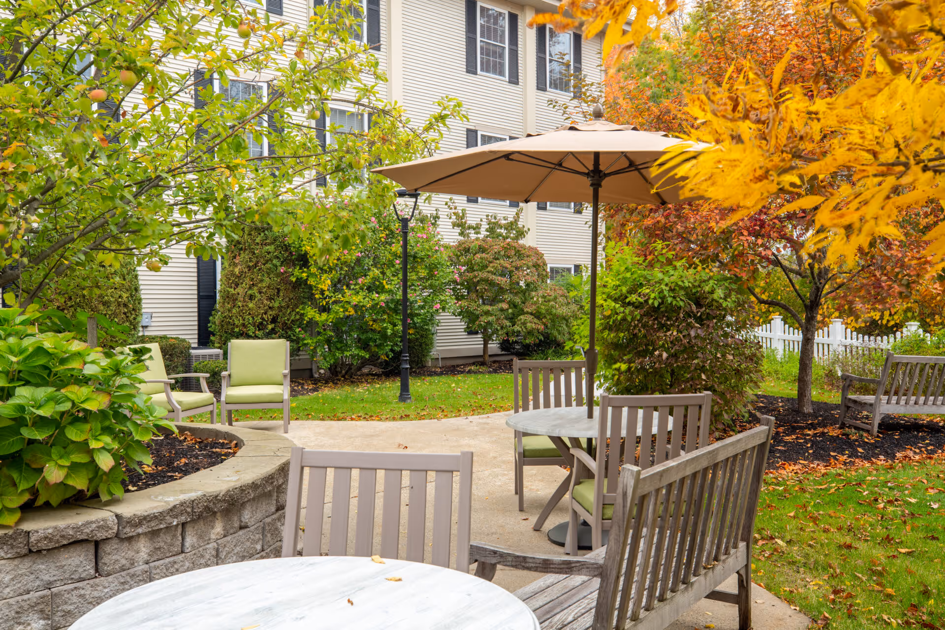 Outdoor seating area in a garden with wooden benches and chairs around round tables, one table shaded by a large beige umbrella. The area is surrounded by green bushes and trees with autumn-colored leaves, next to a beige building with multiple windows.