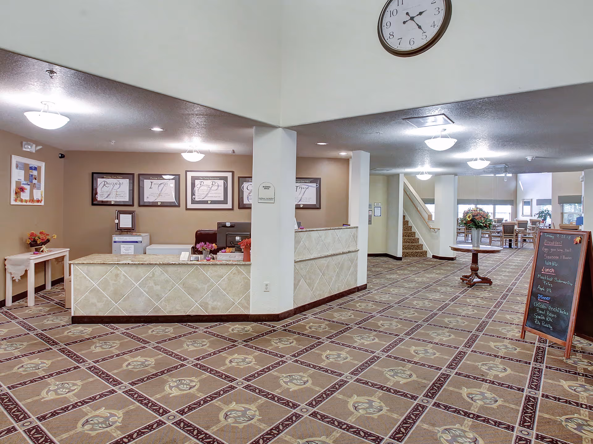 Spacious senior living facility lobby with a reception desk, patterned carpet, a small table with flowers, and a chalkboard sign.