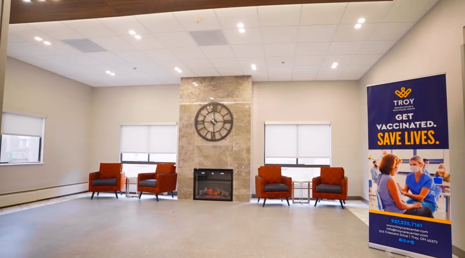 A spacious, well-lit common area with four orange armchairs arranged in pairs on either side of a central fireplace with a large clock above it. Two windows with white blinds are on the back wall. To the right, there is a standing banner promoting vaccination with the text 'Troy Rehabilitation & Healthcare Center, Get Vaccinated. Save Lives.' and contact information.