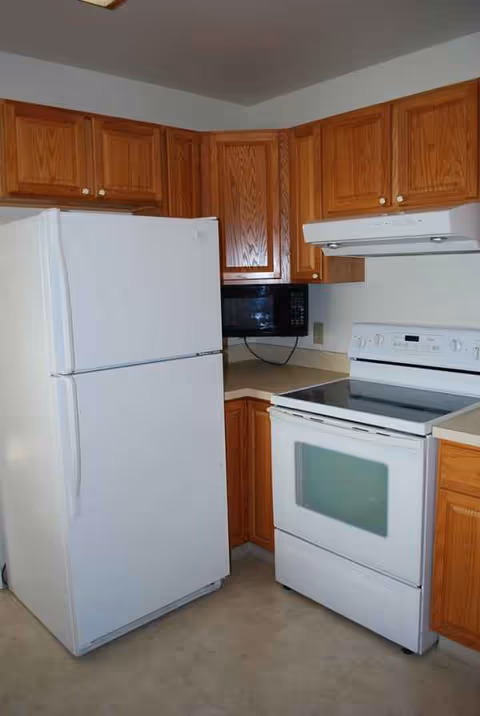 Compact kitchen with white fridge, stove, microwave, and wooden cabinets in a senior living facility.