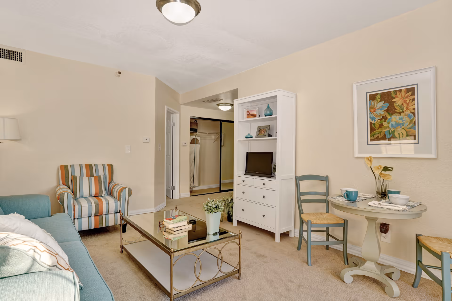 A cozy living room area with a blue sofa, a striped armchair, a glass coffee table with books and a plant, a white shelving unit with decorative items and a small TV, and a small round table set with two chairs, cups, and a vase with flowers. The walls are beige with a floral painting, and there is a doorway leading to a closet area.
