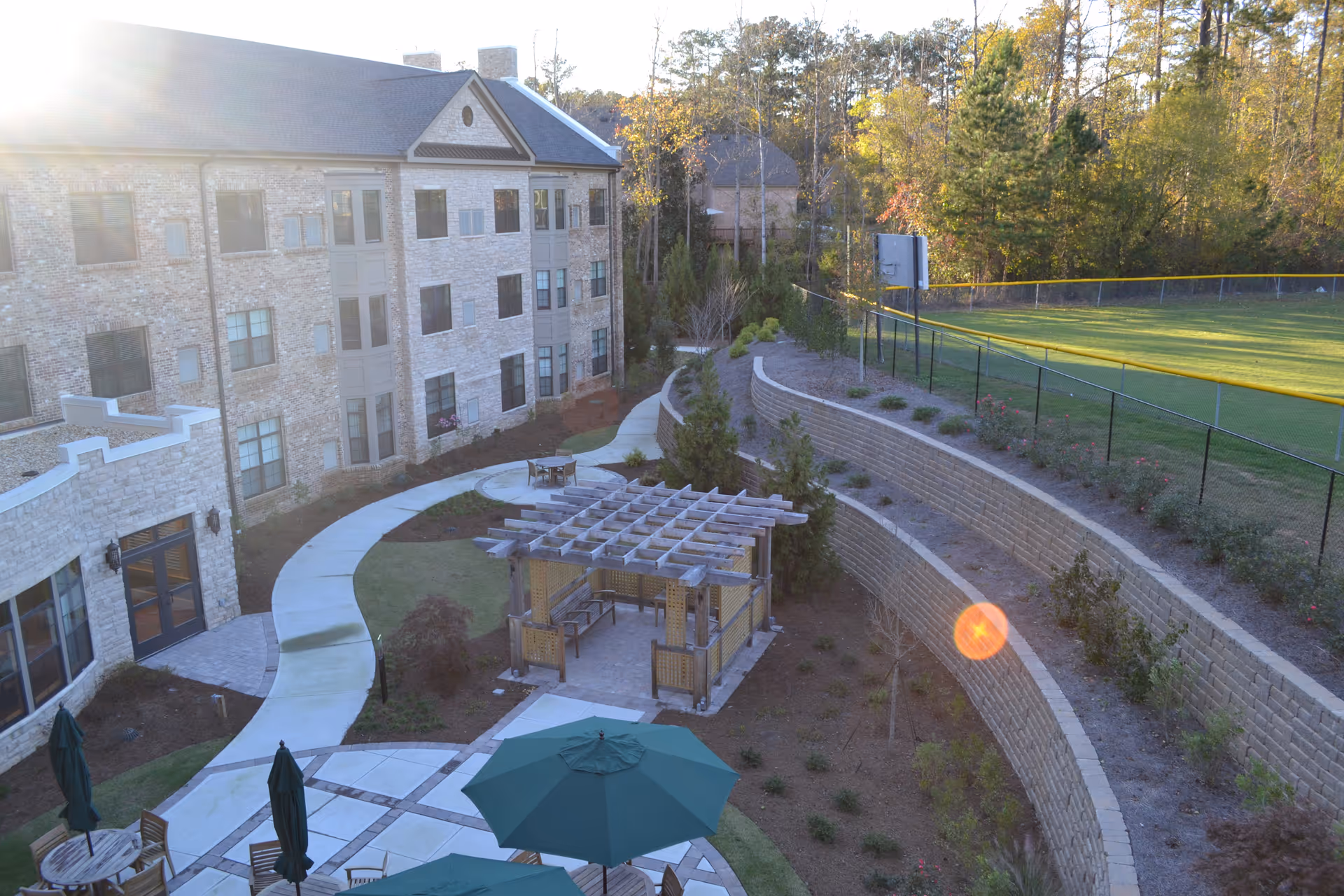 Outdoor courtyard with seating tables, a pergola, winding walkway, and a multi-story building beside a fenced sports field.