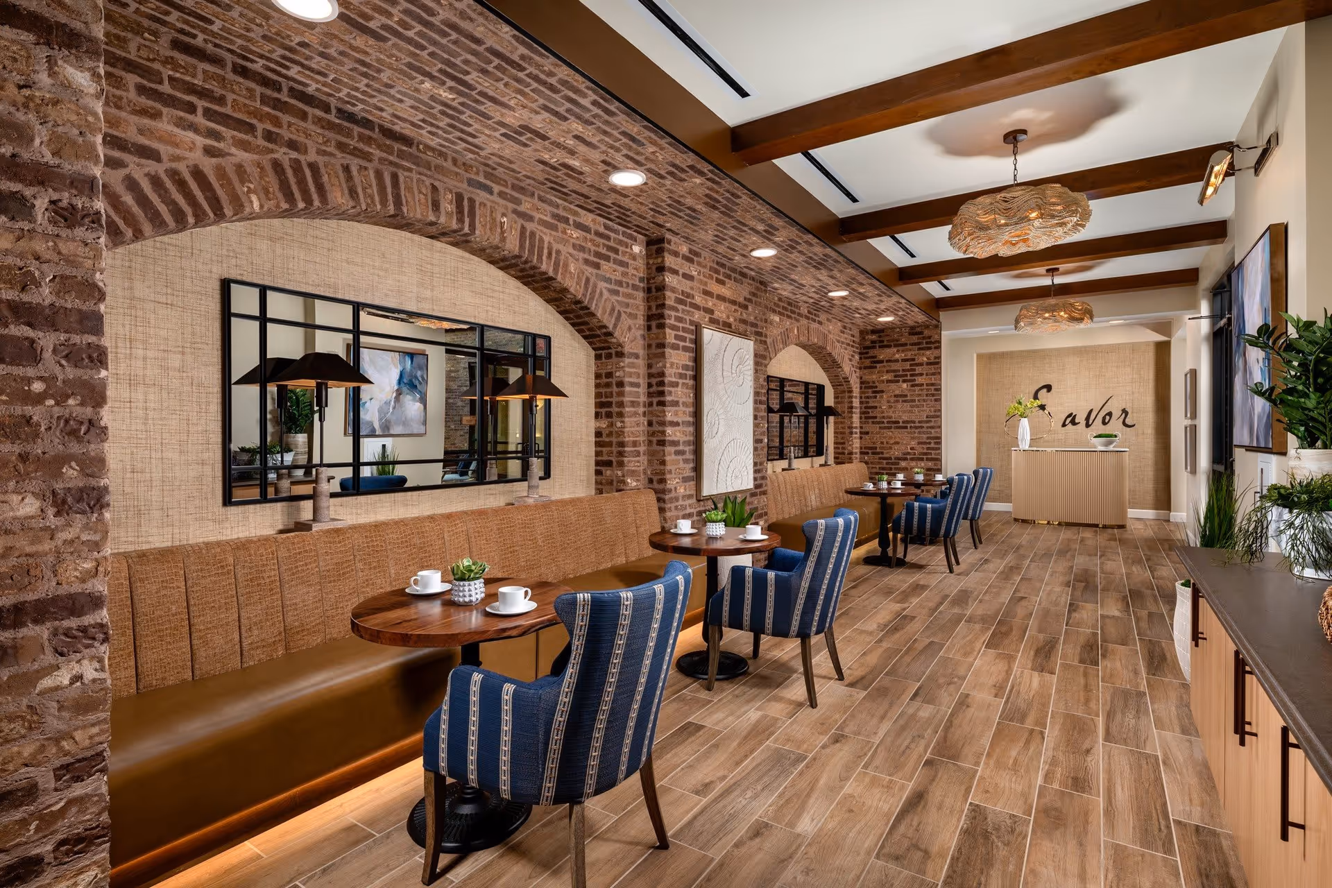 A cozy dining area with wooden tables and blue upholstered chairs. The space features exposed brick arches, a long cushioned bench along the wall, decorative wall art, and pendant lights hanging from a ceiling with wooden beams. There are plants and cups on the tables, and a sign that reads 'Savor' on the far wall.