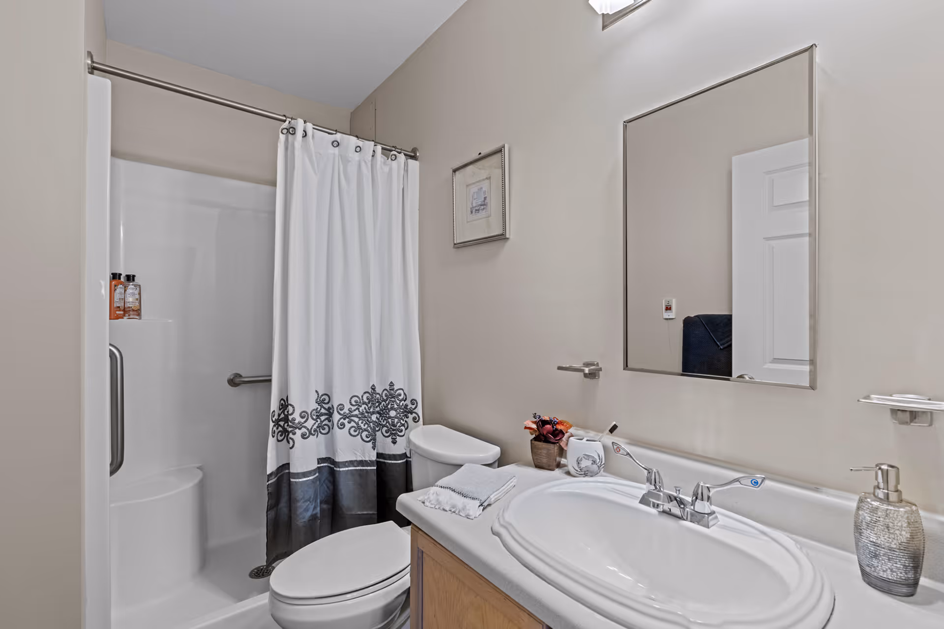 A clean bathroom featuring a white sink with silver faucet, a large rectangular mirror above it, a toilet, and a shower with a white curtain decorated with a black pattern. The walls are painted beige, and there is a small framed picture hanging on the wall.