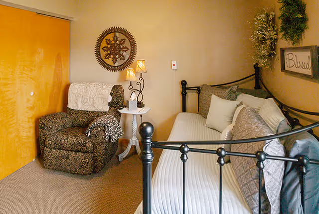 A cozy room with a black metal daybed adorned with multiple pillows and a white quilted cover. Next to the bed is a small white side table with a decorative lamp and a small box. To the left, there is a patterned armchair with a white throw draped over the back. The wall behind features a round decorative wall hanging and a framed sign that reads 'Blessed' with some greenery above it. The room has beige walls and carpeted floor.