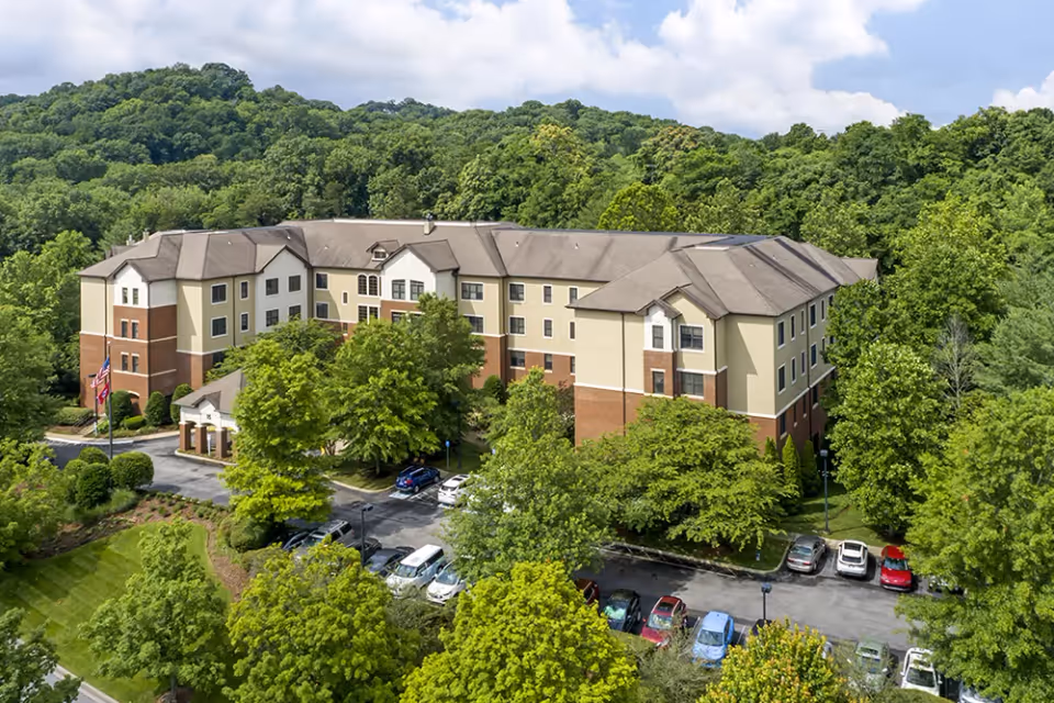 Aerial view of a multi-story senior living facility building surrounded by lush green trees and hills under a partly cloudy sky. Several cars are parked in the parking lot in front of the building.