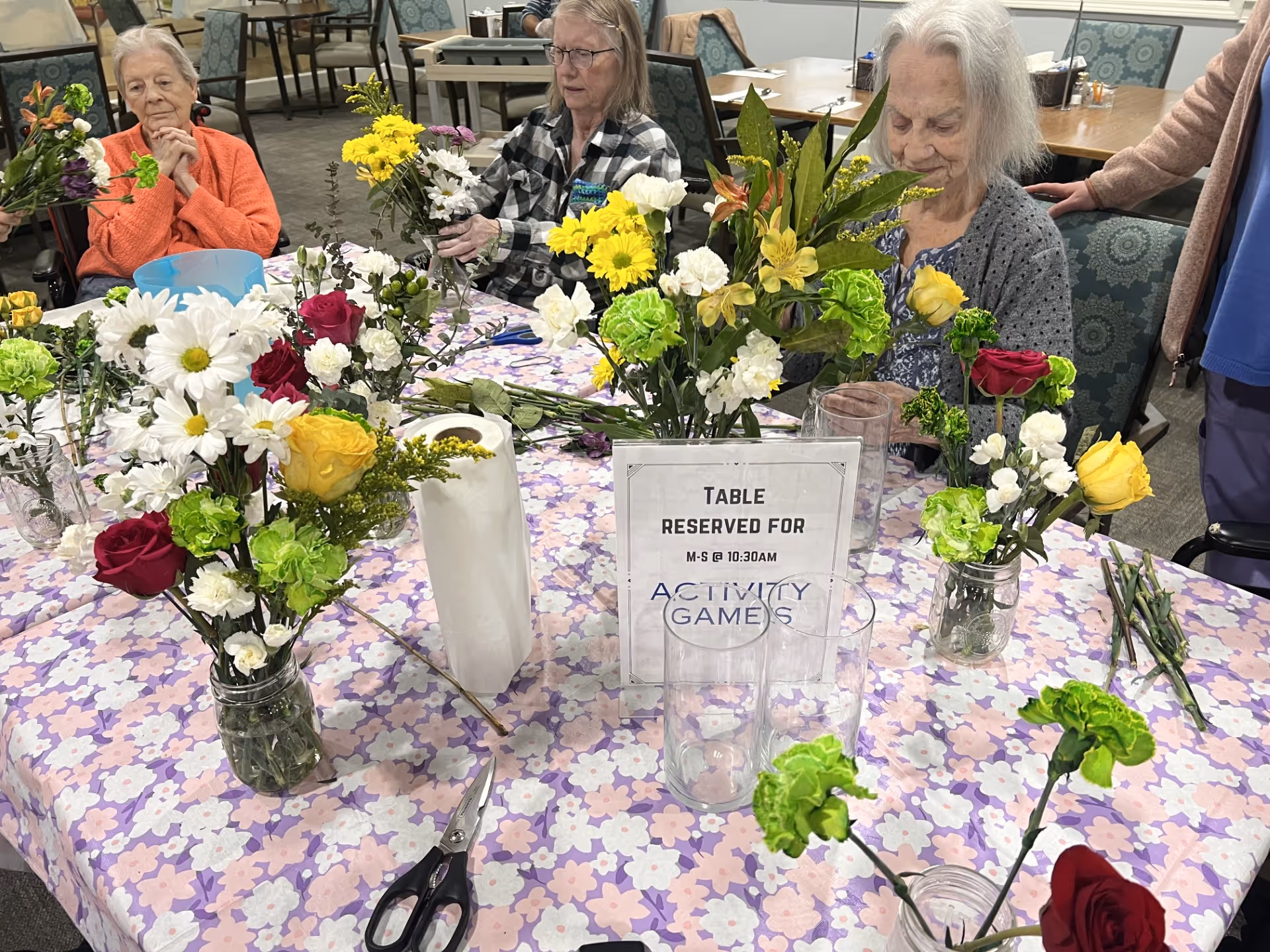 Three elderly women sitting around a table covered with a floral tablecloth, arranging colorful flowers in glass jars. The table has a sign that reads 'TABLE RESERVED FOR M-S @ 10:30AM ACTIVITY GAMES'. Scissors and paper towels are also on the table. The setting appears to be a communal activity room with chairs and tables in the background.