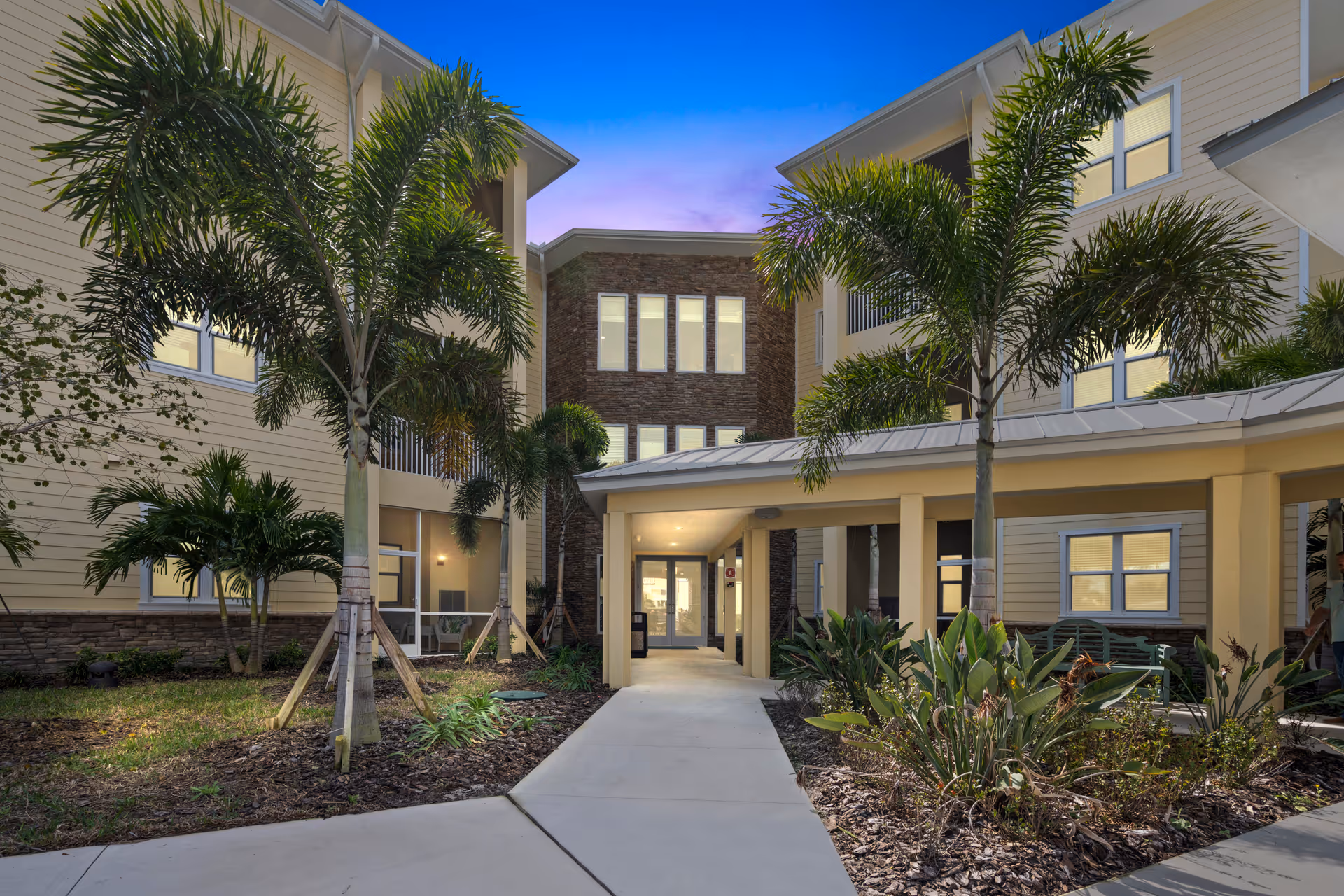 Exterior view of The Residences at Zon Beachside showing a walkway leading to the entrance of a multi-story building with palm trees and landscaped plants on either side under a twilight sky.