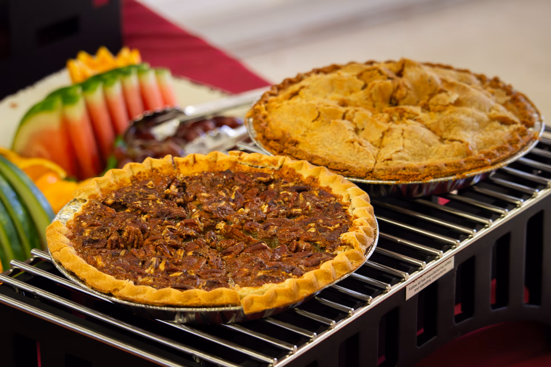 Two pies (pecan and apple) displayed on a buffet rack with sliced watermelon and other fruit in the background.
