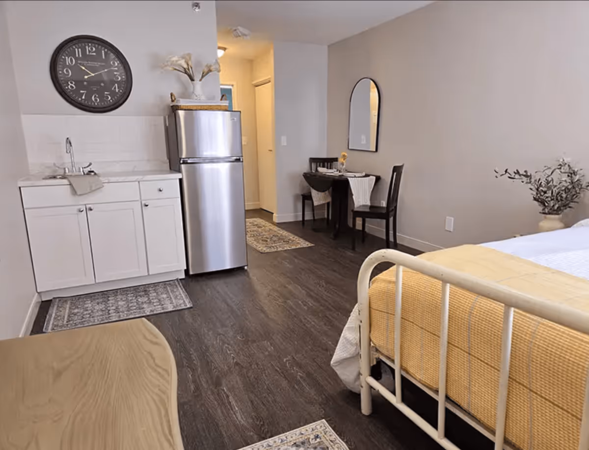 A studio apartment interior featuring a small kitchenette with a sink, white cabinets, and a stainless steel refrigerator. A round dining table with two chairs is set against the wall with a mirror above it. A bed with a yellow blanket is partially visible on the right side. The room has dark wood flooring and neutral-colored walls.