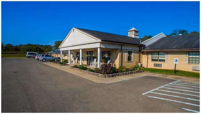 Exterior view of a single-story healthcare facility building with a covered entrance supported by white columns. Several cars are parked along the driveway, and there is a handicapped parking space visible in the foreground. The sky is clear and blue.
