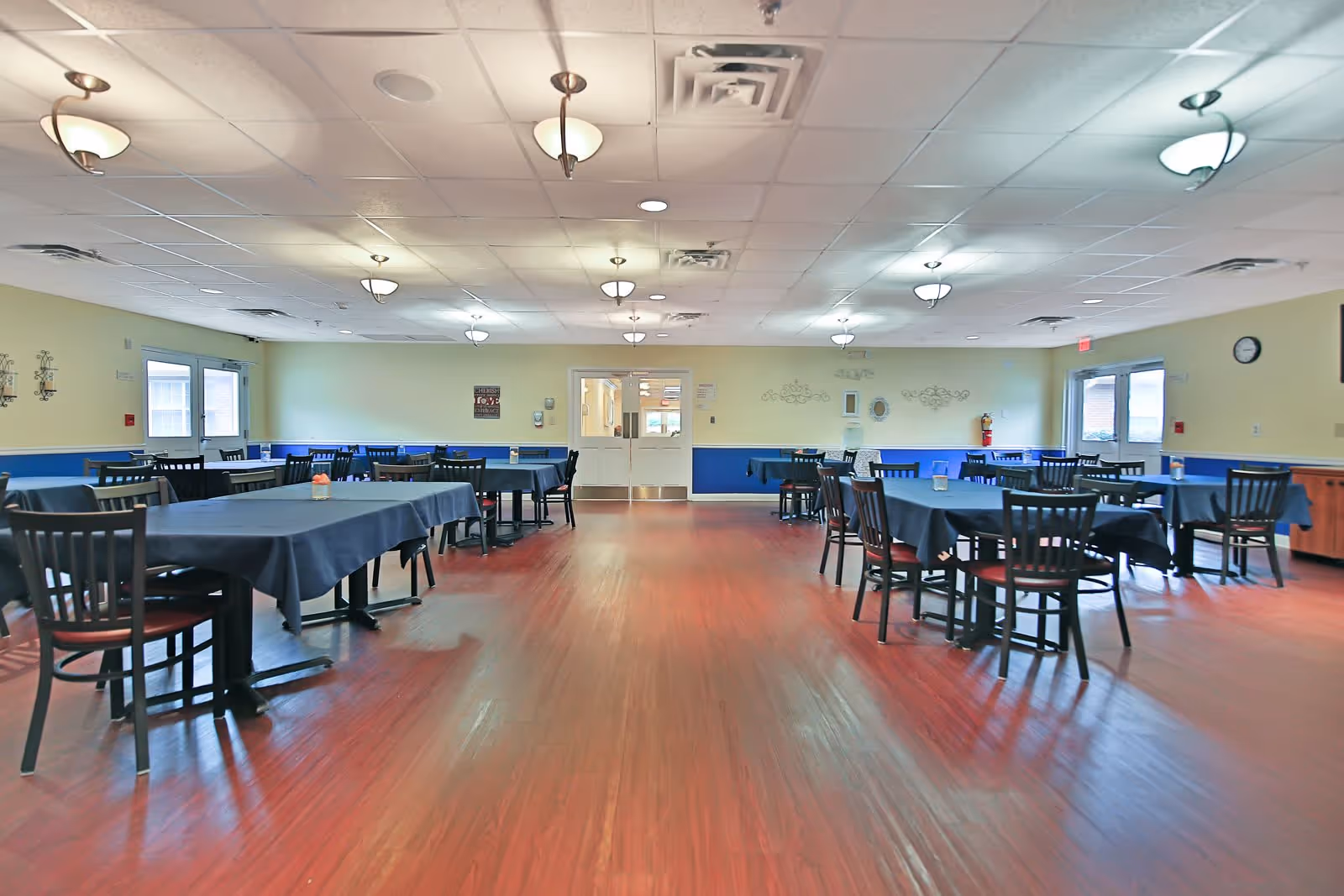 A spacious dining room with multiple tables covered in dark blue tablecloths and surrounded by black chairs with red cushions. The room has wooden flooring, pale yellow walls with a blue wainscot, and ceiling lights evenly spaced. There are double doors at the far end and windows on the left and right walls.