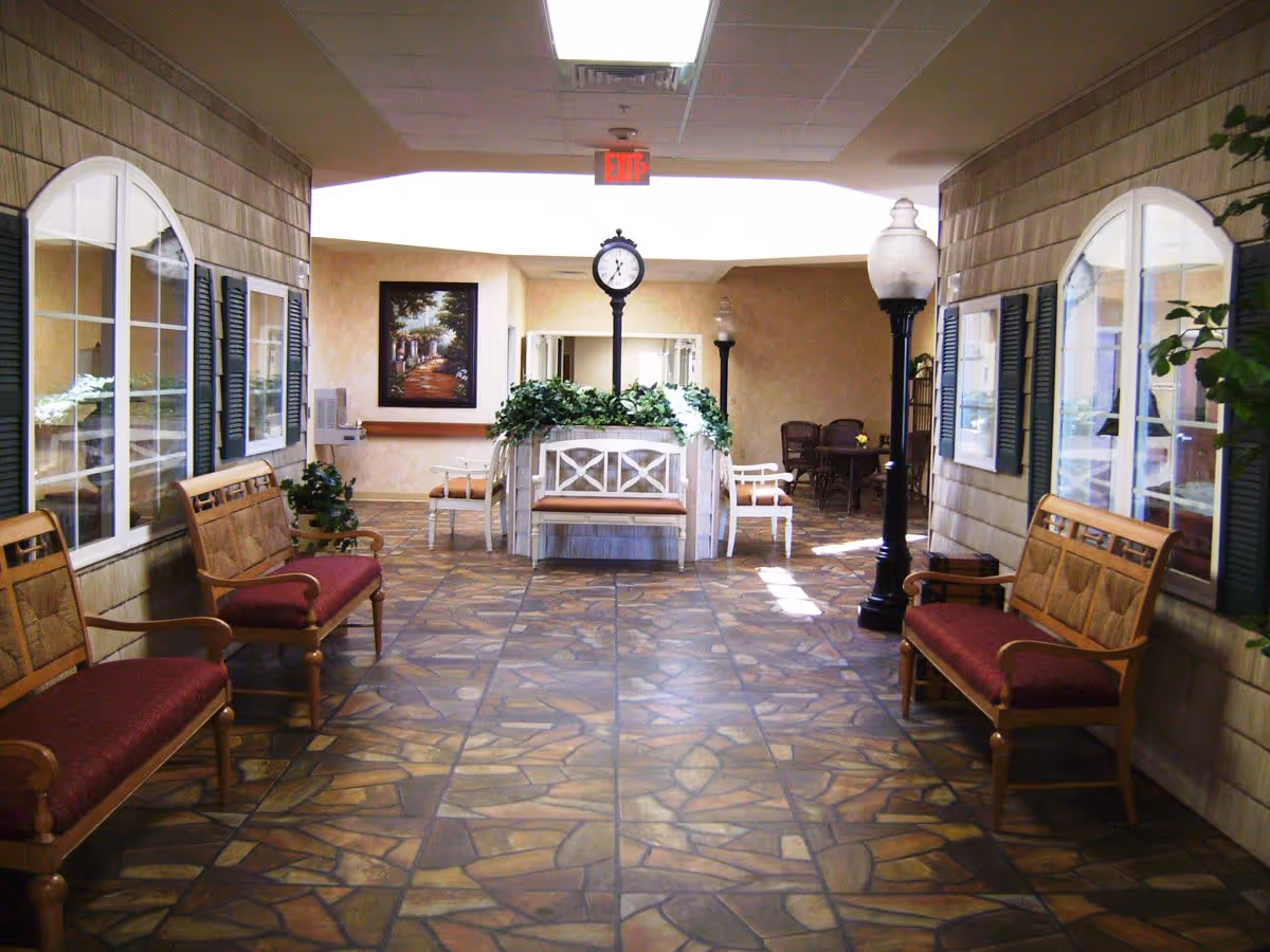 Indoor hallway area with patterned tile floor, wooden benches with red cushions along both sides, decorative street lamps, a clock on a post surrounded by plants, and a framed painting on the far wall.