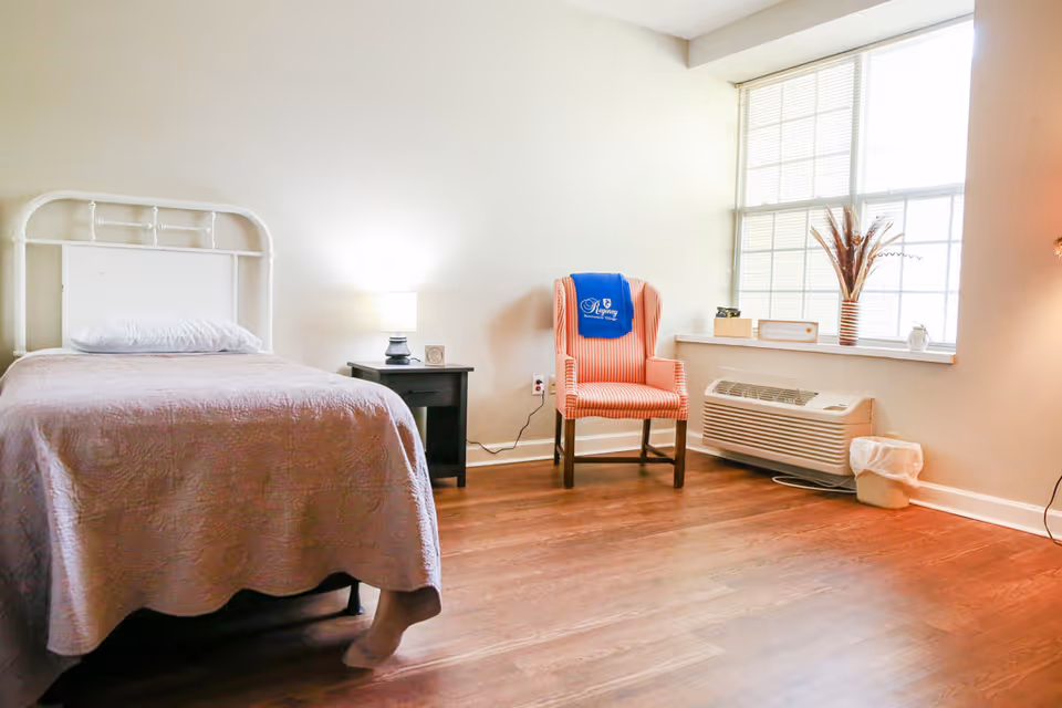 A simple bedroom with a single bed covered in a light pink quilt, a pillow, a black nightstand with a lamp, and a striped armchair with a blue Regency Retirement Village of Jackson blanket draped over the back. The room has wooden flooring, a large window with blinds, a white air conditioning unit below the window, and a small trash bin beside it.
