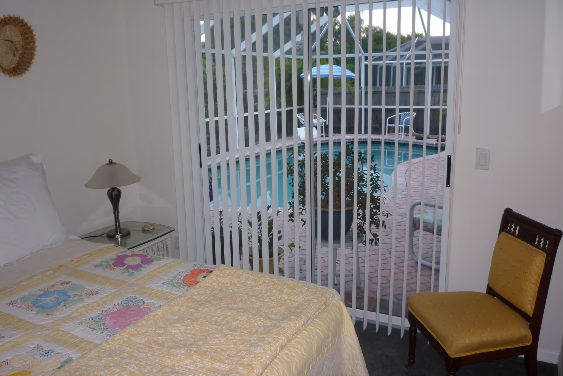 A bedroom with a bed covered in a yellow floral quilt, a glass-top bedside table with a lamp, a yellow upholstered chair, and vertical blinds covering a sliding glass door that opens to a patio with a swimming pool and outdoor chairs.