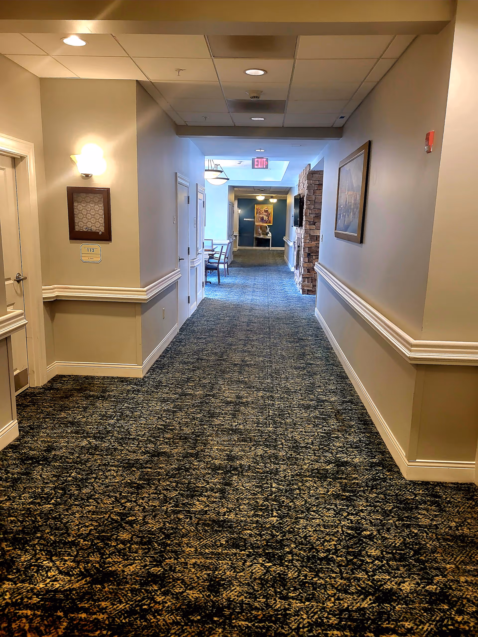 Carpeted interior hallway in a senior living facility with doors along the sides and seating visible at the far end.