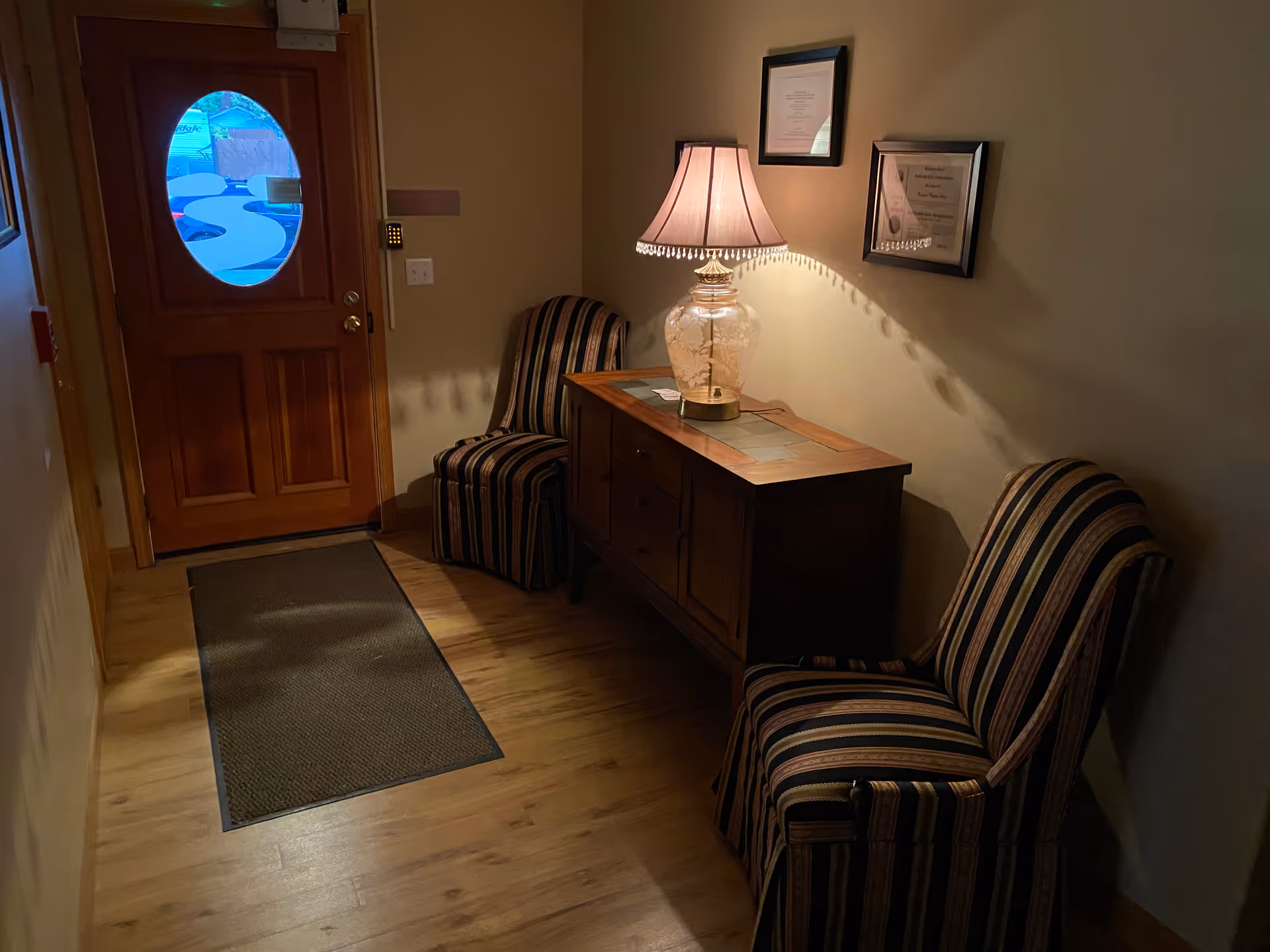 A small entryway with a wooden door featuring an oval window. Inside, there are two striped upholstered chairs on either side of a wooden console table with drawers. A decorative lamp with a beige shade is on the table, casting light and shadows on the wall. Two framed certificates or documents hang above the table. The floor is wooden with a dark floor mat near the door.