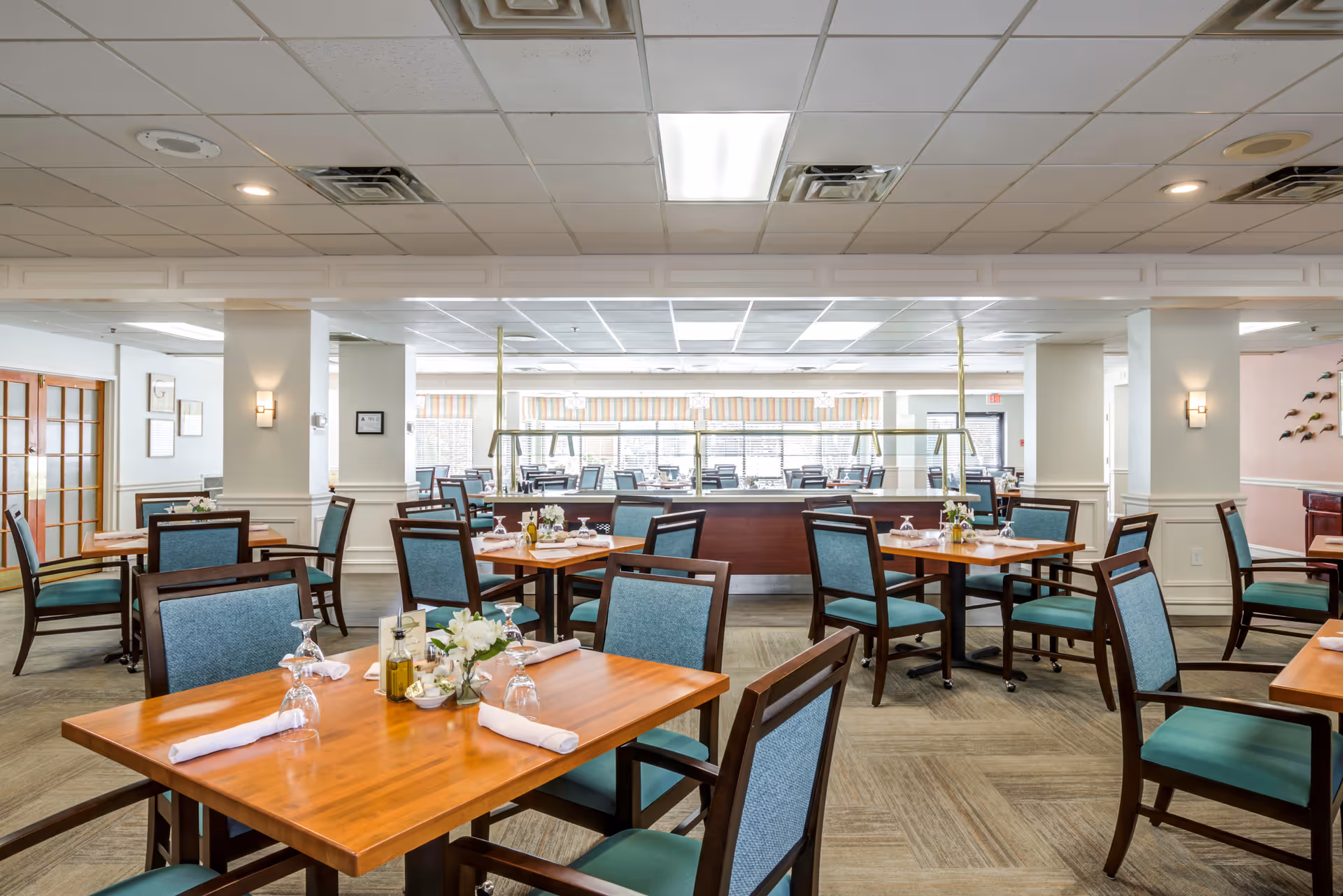 Spacious dining room with multiple wooden tables and chairs with blue upholstery. Tables are set with white napkins, upside-down wine glasses, and small flower arrangements. The room has a light-colored carpet, white walls with decorative molding, and large windows in the background letting in natural light.