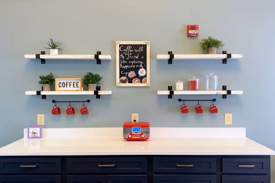 A coffee station with a white countertop and black cabinets below. Above the counter are two sets of wooden shelves mounted on a light blue wall. The shelves hold small potted plants, jars, and a sign that says 'COFFEE'. Red coffee mugs hang from rods under the shelves. A framed chalkboard sign in the center reads 'Coffee with a friend is like capturing happiness in a cup'. A small red radio sits on the counter.