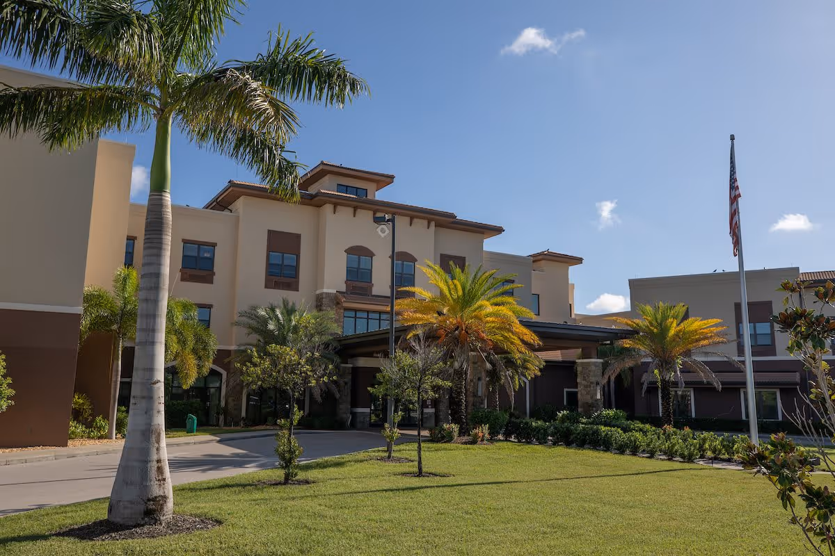 Exterior view of Keystone Place at Beachwalk senior living facility with a well-maintained lawn, palm trees, and an American flag on a flagpole under a clear blue sky.
