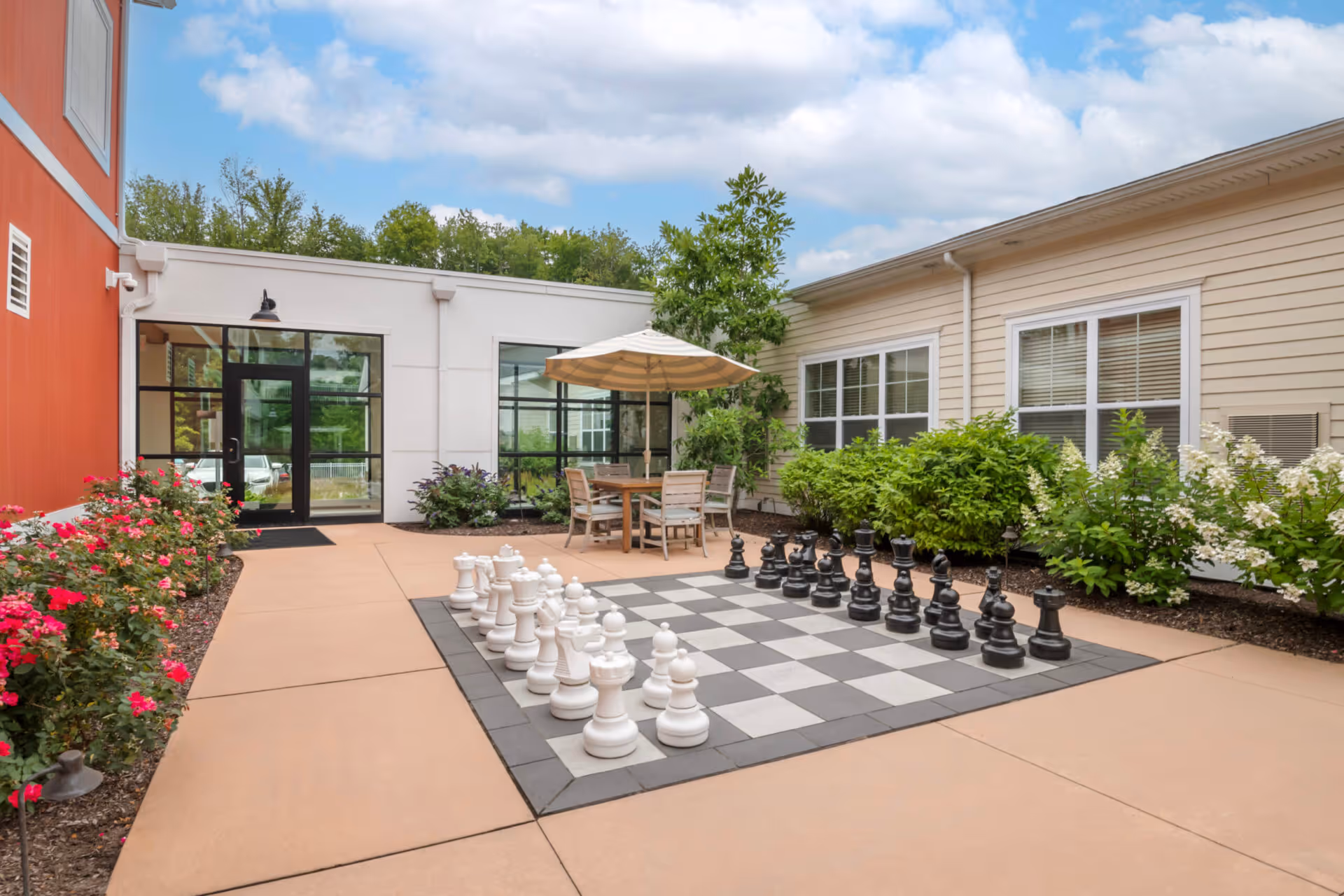 Outdoor courtyard area at Church Hill Village featuring a large outdoor chessboard with oversized chess pieces, a table with chairs and an umbrella, surrounded by bushes and flowers, with building walls and windows in the background under a partly cloudy sky.