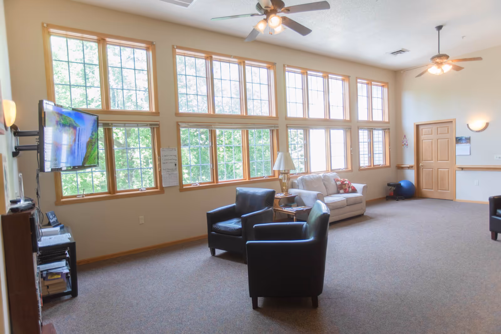 Sunlit senior living lounge with tall multi-pane windows, two black armchairs, a beige sofa, and a wall-mounted TV.
