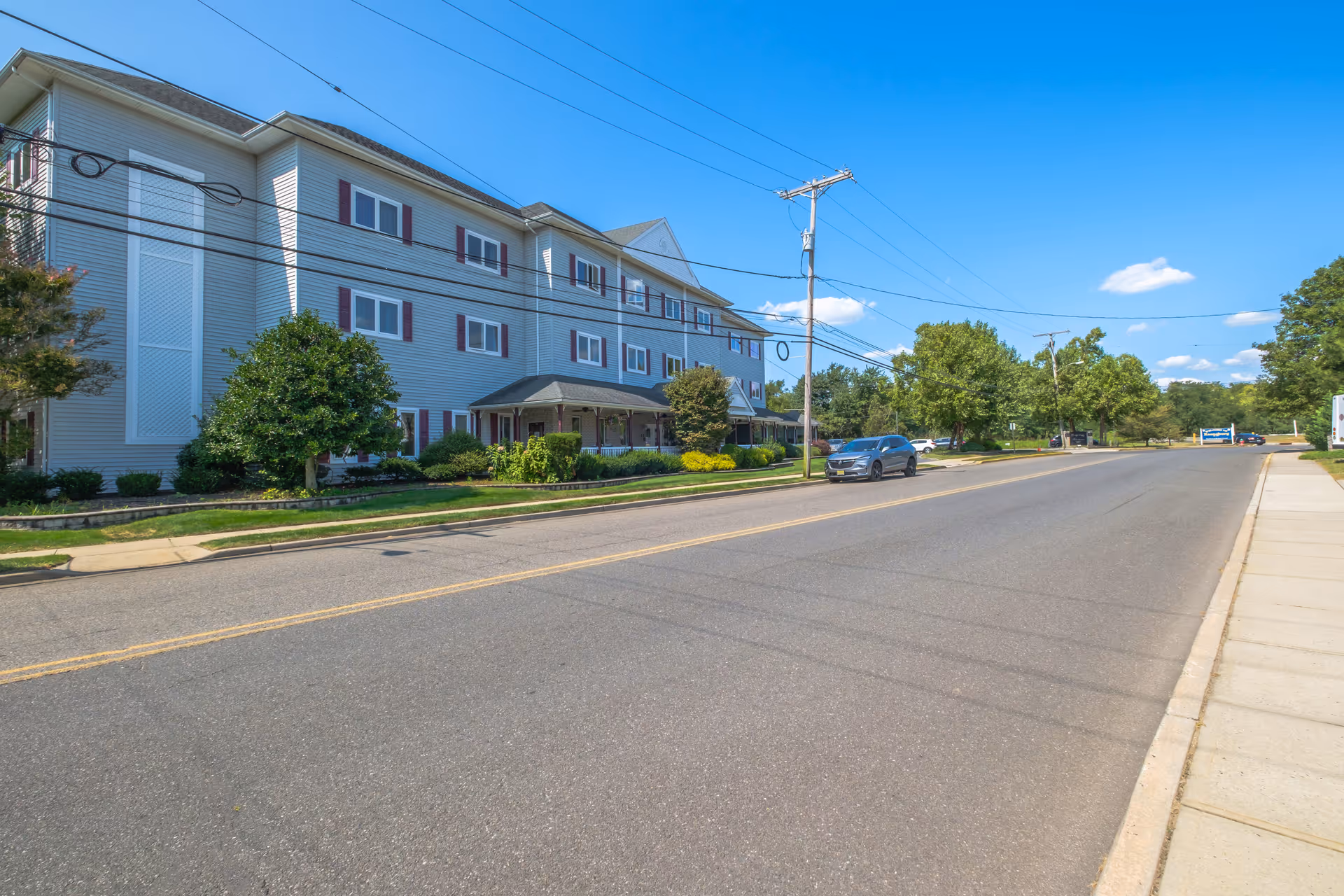 Three-story light-gray residential care building along a quiet suburban street with a parked car, trees, and a clear blue sky.