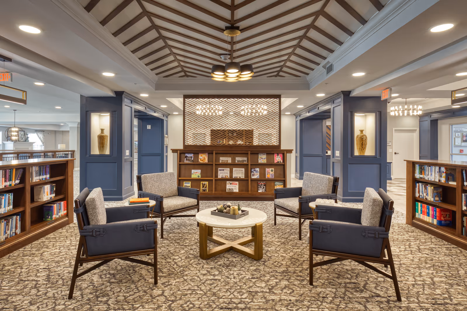 A cozy and well-lit library or reading area with four cushioned armchairs arranged around a round coffee table. The space features built-in bookshelves filled with books and magazines, decorative vases in wall niches, and a patterned carpet. The ceiling has a wooden beam design with modern light fixtures.