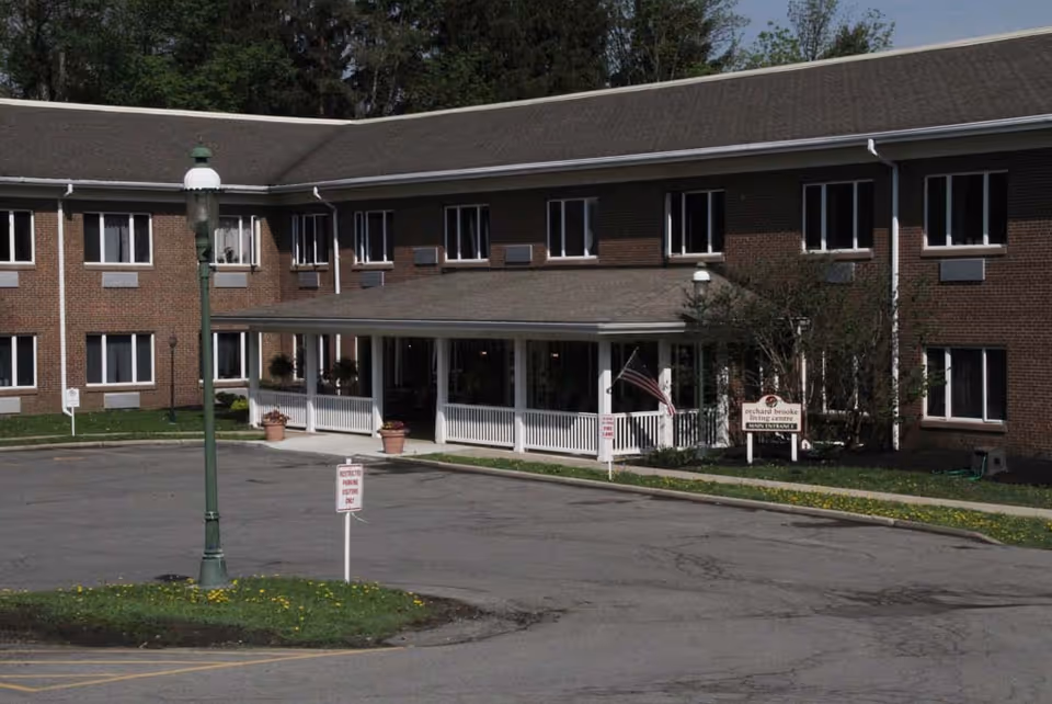 Exterior view of Orchard Brooke Living Center, a two-story brick building with multiple windows. The main entrance features a covered porch with white railings and potted plants. An American flag is displayed near the entrance. There is a parking area in front with signs indicating parking restrictions. Trees and greenery surround the building.