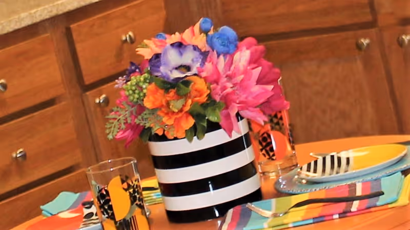 A close-up view of a dining table set with colorful striped napkins, plates, and glasses. In the center of the table is a black and white striped vase holding a vibrant bouquet of flowers in various colors including pink, orange, purple, and blue. Wooden cabinets with silver knobs are visible in the background.