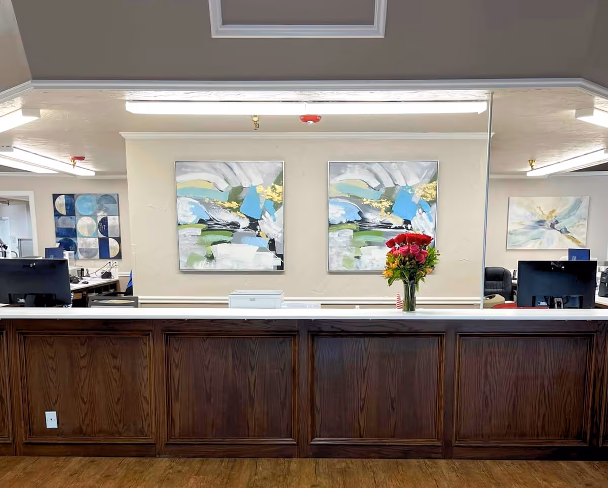 Reception desk area with a wooden front panel and a white countertop. Behind the desk, there is a large mirror reflecting two abstract paintings with blue, green, white, and gold colors. A vase with red and yellow flowers is placed on the counter. In the background, office desks with computer monitors and additional abstract artwork on the walls are visible.