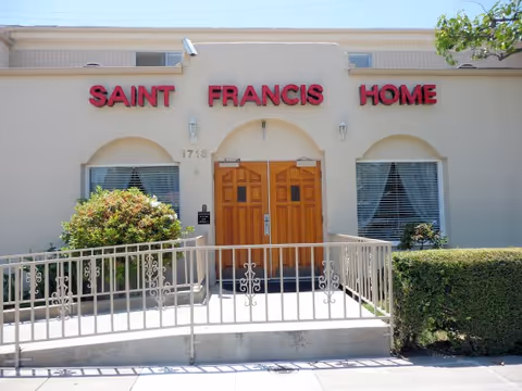 Front exterior view of Saint Francis Home building with beige walls, two wooden double doors, windows with blinds on either side, and bushes in front. A metal railing leads up to the entrance.