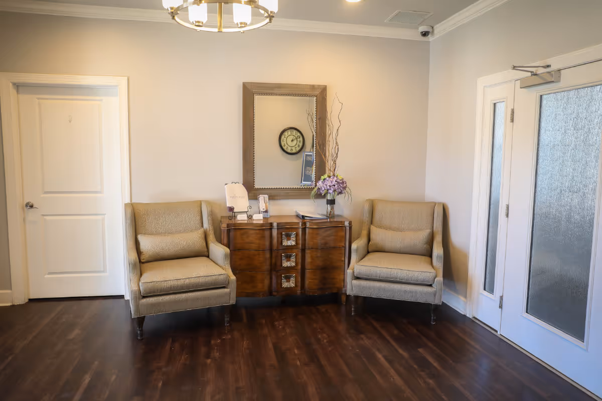 A cozy seating area in a memory care community with two beige armchairs on either side of a wooden cabinet. A large mirror hangs above the cabinet, reflecting a wall clock. The room has light-colored walls, dark wooden flooring, a white door on the left, and a glass-paneled door on the right.