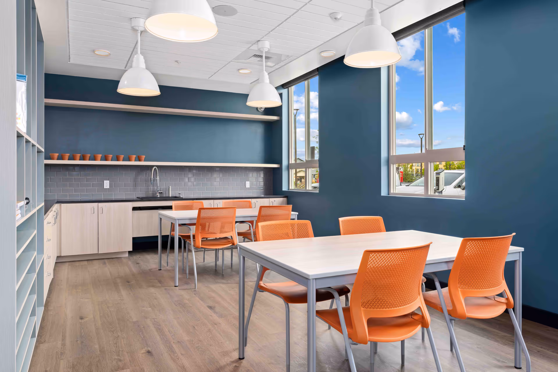 A modern room with blue walls and large windows letting in natural light. The room features two white tables with orange chairs around them. There are white pendant lights hanging from the ceiling. Along one wall is a countertop with a sink, light wood cabinets, and open shelves holding small terracotta pots.