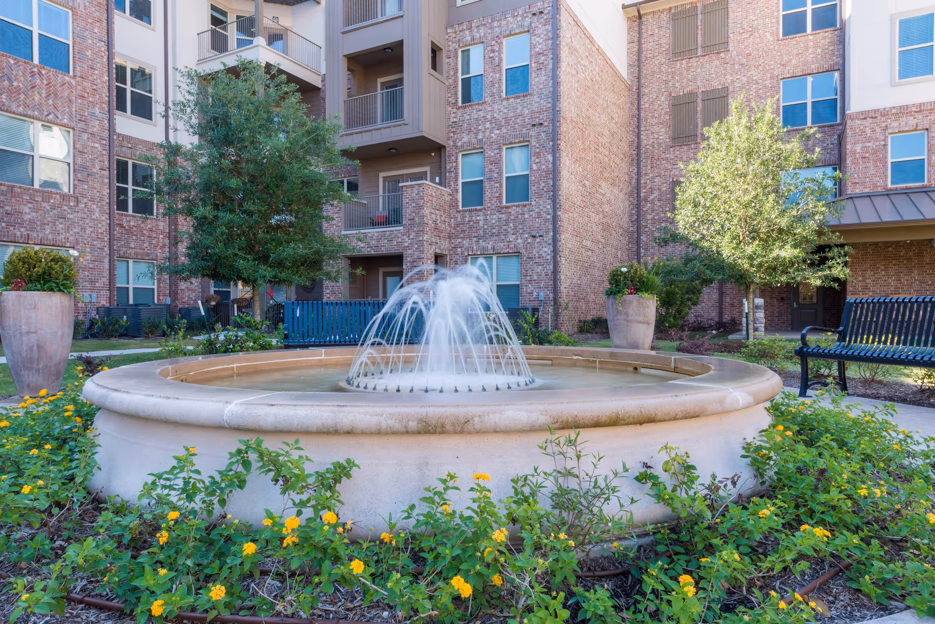 Outdoor courtyard area at Watercrest at Katy featuring a circular water fountain surrounded by green plants with yellow flowers, two large planters, a black metal bench, and a multi-story brick building in the background.