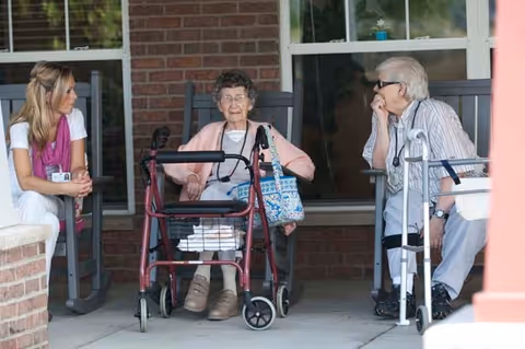 Two elderly women sitting on rocking chairs on a porch, each with a walker in front of them, engaged in conversation with a younger woman sitting nearby.