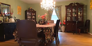 A dining room with a wooden table covered by a floral tablecloth surrounded by six dark upholstered chairs. The room features a chandelier hanging from the ceiling, a large window with floral curtains, and two wooden china cabinets filled with decorative items. The walls are painted light green and decorated with framed artwork.