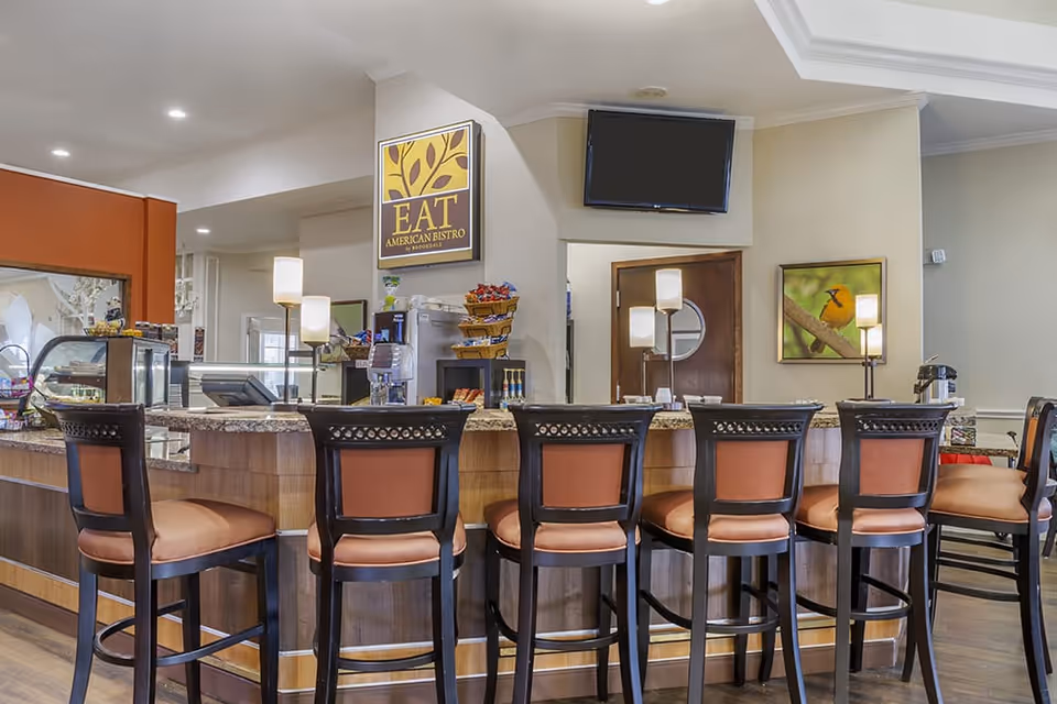 Interior view of a dining area with a counter and six high chairs with orange cushions. Behind the counter are lamps, a coffee machine, baskets with snacks, and a sign that reads 'EAT American Bistro'. A flat-screen TV is mounted on the wall above the counter, and a framed picture of a bird on a branch is visible on the right wall.